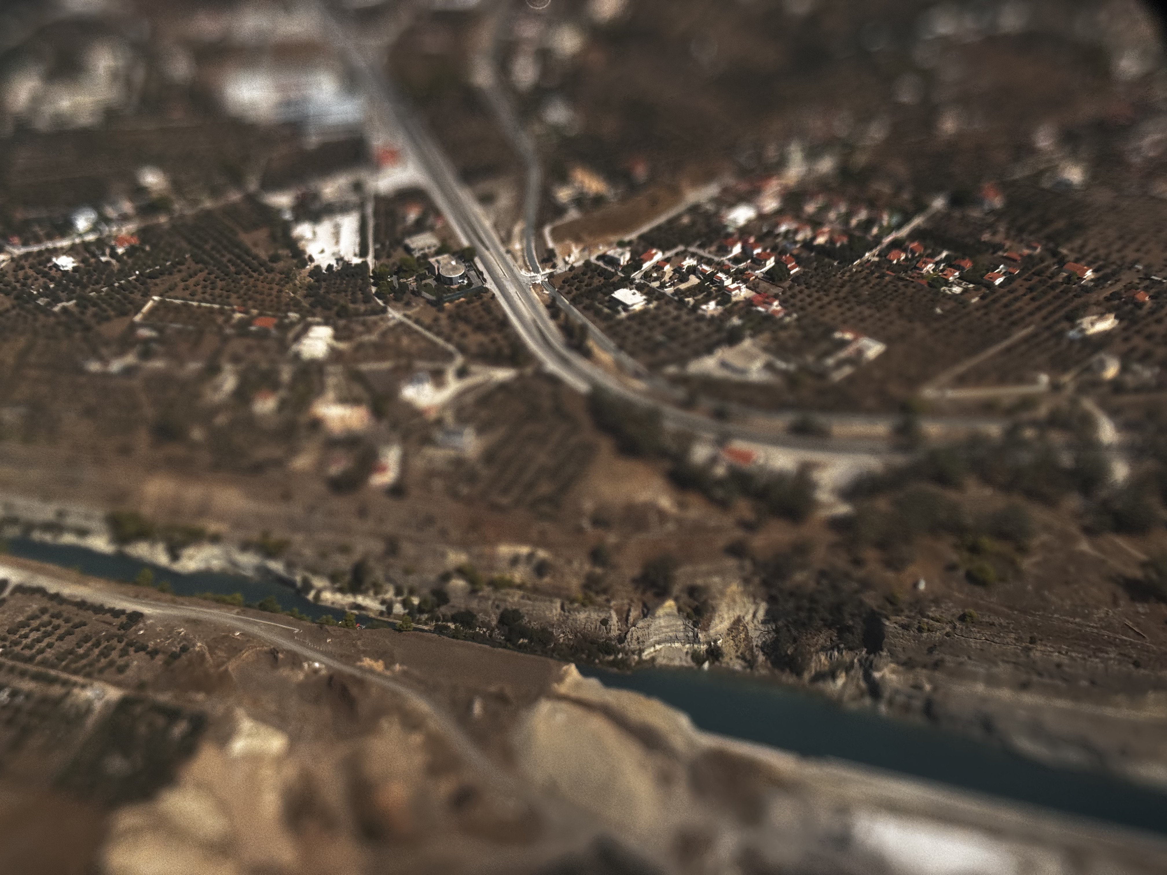Aerial view looking down at the Corinth Canal and the land around it — a highway, clusters of white houses with terracotta roofs, and olive groves — with the top and bottom of the scene softly blurred so it reads like a miniature model