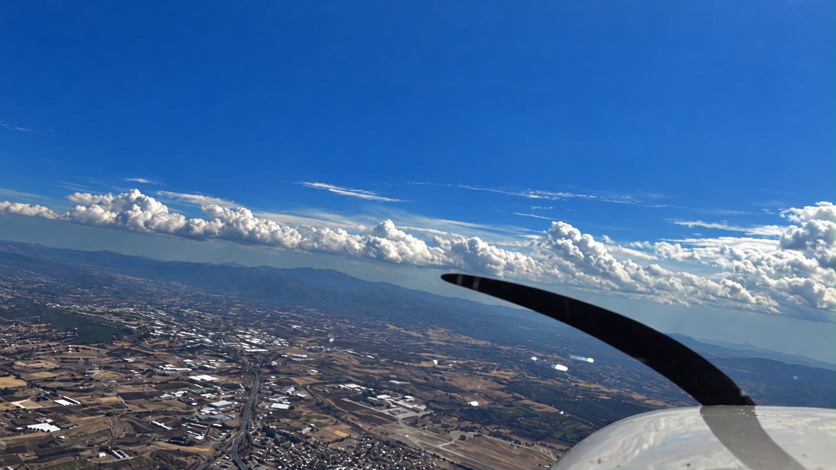 View from a small aircraft: the black propeller blade and grey nosecone in the lower right, above a patchwork of farmland and greenhouse roofs with a bank of white cloud along the horizon