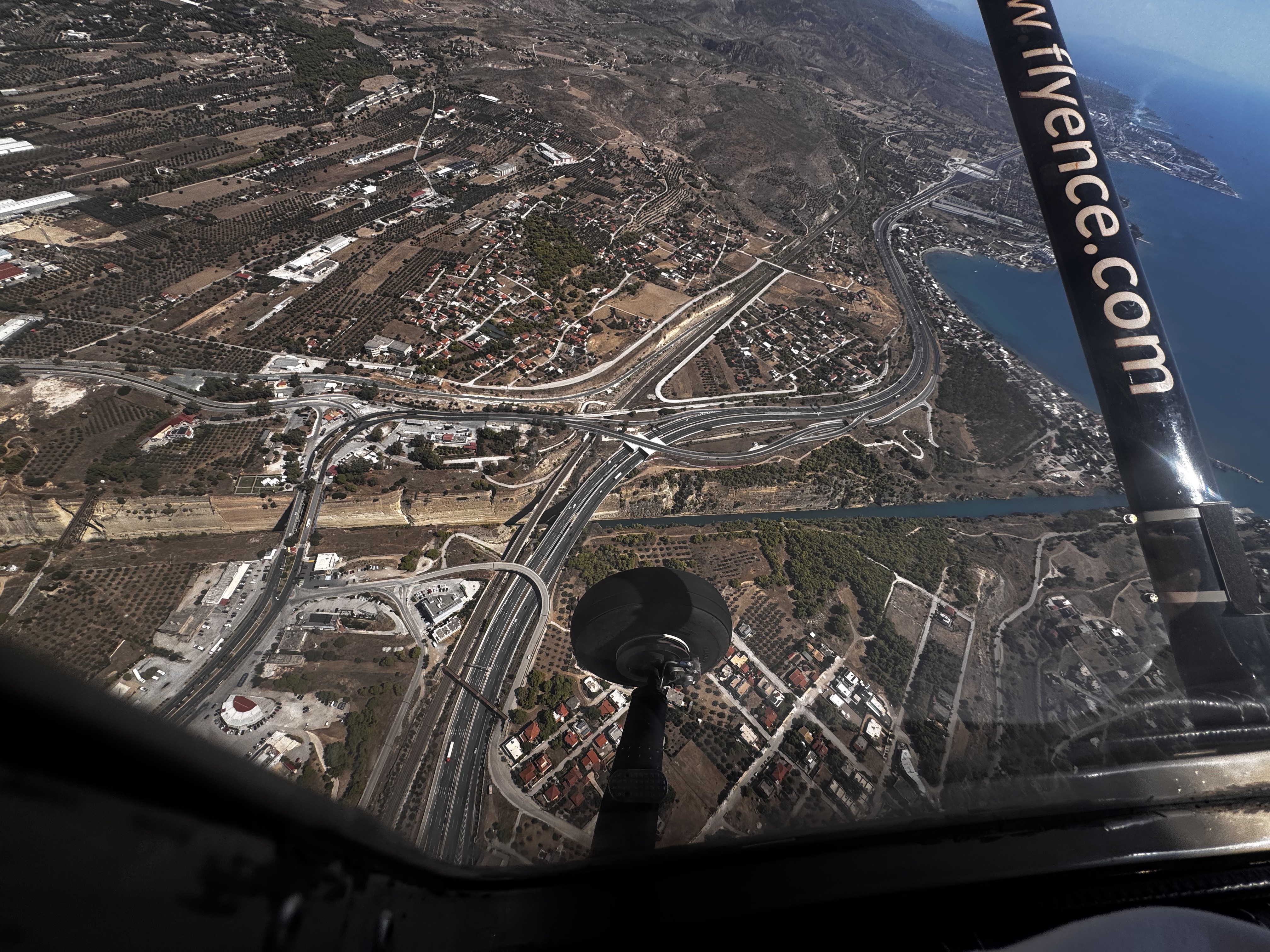 Aerial view from inside a small aircraft, the black landing gear wheel and strut in the foreground, above a highway interchange, the straight cut of the Corinth Canal and the blue coastline beyond